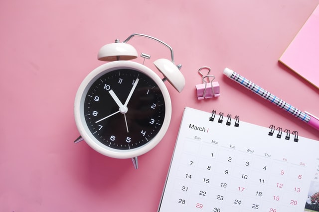 Picture showing a white alarm clock and a planner on a pink background. 