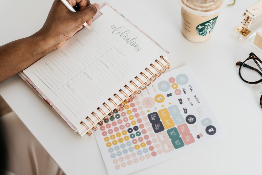 Image showing a hand holding a pen above a planner. Below the planner is a sheet of colorful stickers.