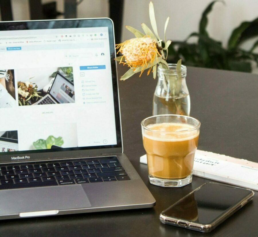 Image showing a desk with an open laptop. On the desk you can also see a glass filled with coffee, a cellphone and a vase with a flower.