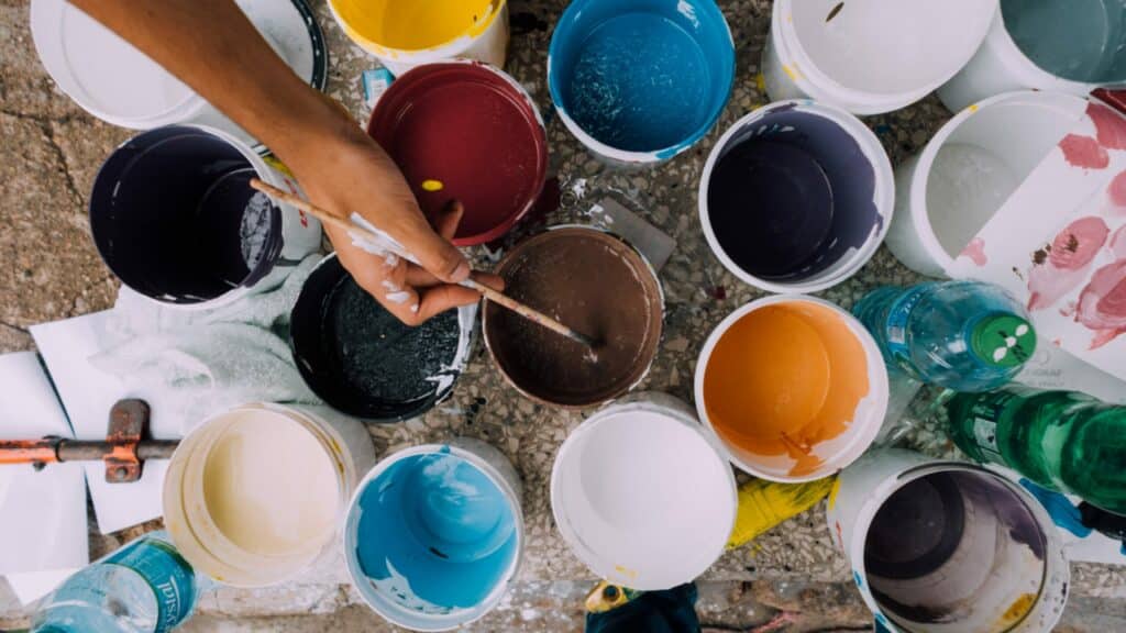 Different cups of paint are seen from above. You also see a hand holding a brush being dipped into one of the paint cups.