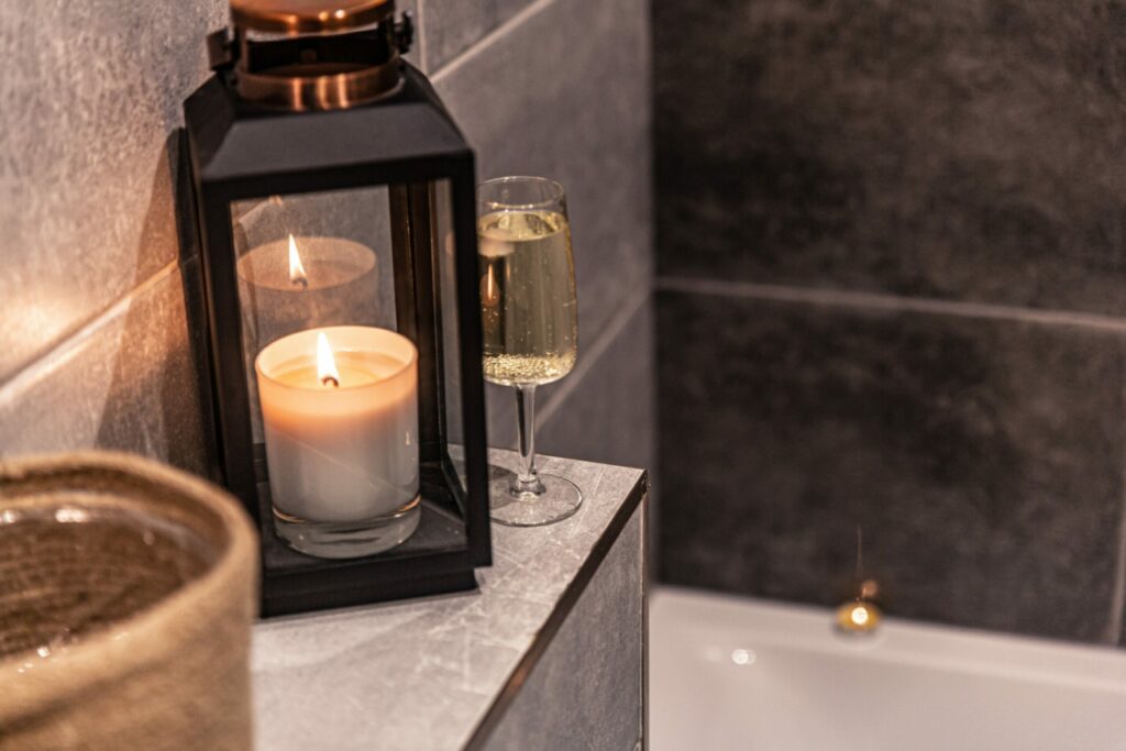 A lit lantern and a glass with bubbly liquid are on a shelf above a bathtub.